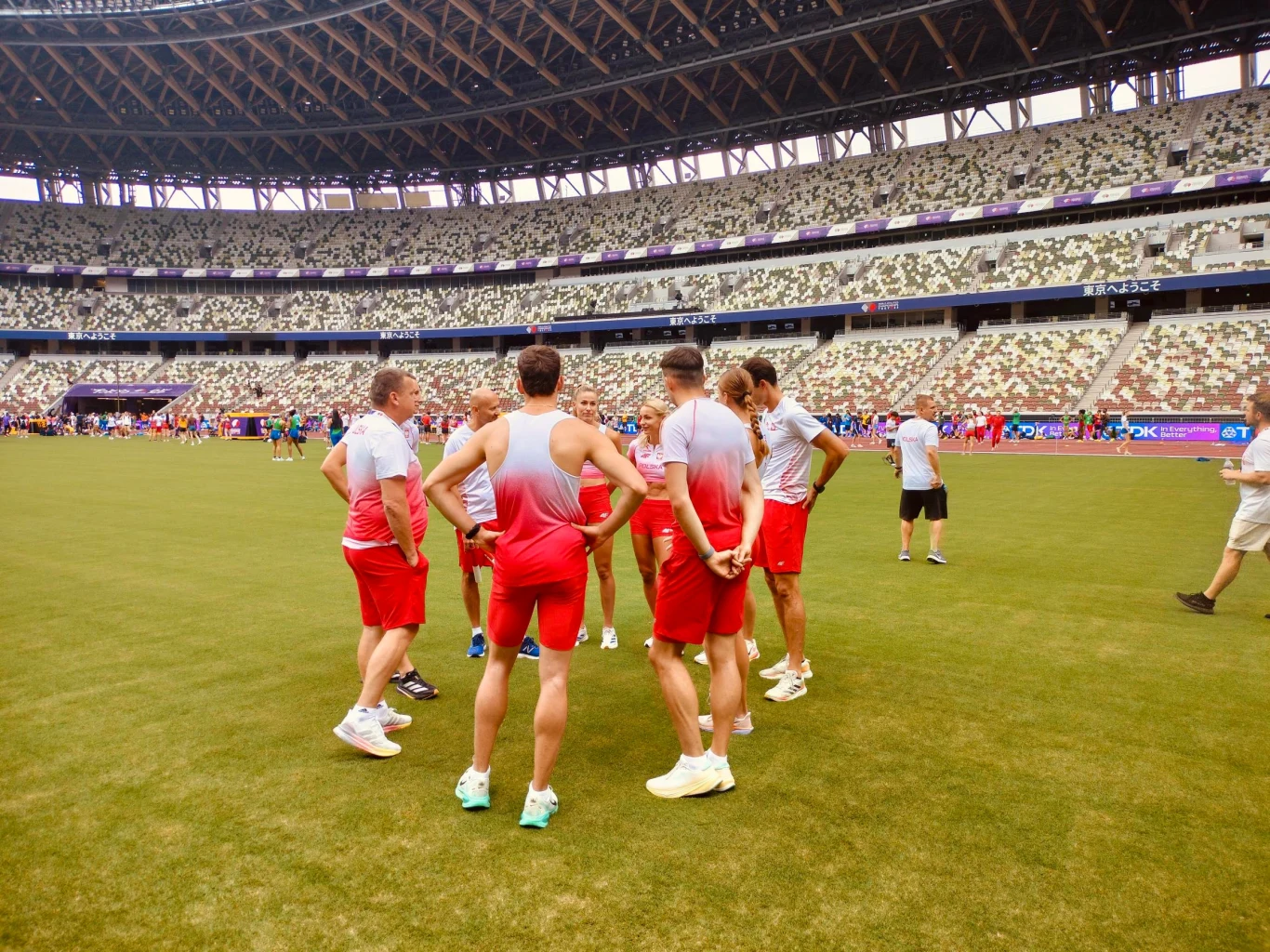 Zebranie polskiej sztafety mieszanej 4x400 m na środku stadionu w Tokio Grupa sportowców w czerwono-białych strojach stoi na murawie stadionu, omawiając strategię przed zawodami, wokół pustych trybun, zdominowanych przez geometryczne wzory siedzeń i zadaszenie.