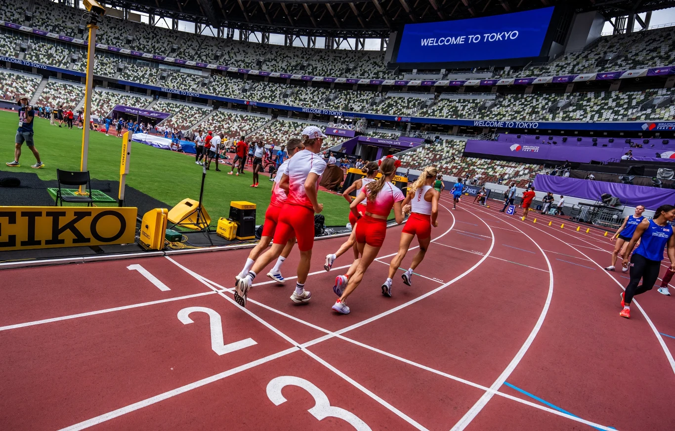 Polska kadra na treningu przed lekkoatletycznymi MŚ w Tokio Grupa sportowców ubranych w czerwone stroje biegnie po bieżni stadionu lekkoatletycznego podczas zawodów, wokół widoczni sędziowie i inni uczestnicy, w tle puste trybuny i duży ekran z napisem 'WELCOME TO TOKYO'.
