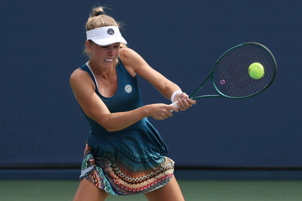 Magdalena Fręch Tenisistka w dynamicznej pozie podczas uderzenia piłki rakietą, ubrana w sportową sukienkę i czapkę z daszkiem, na tle niebieskiego kortu.