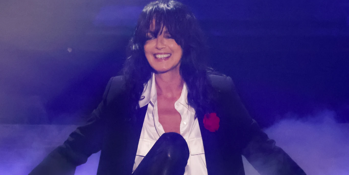 Nena Kobieta z ciemnymi włosami ubrana w biały rozpięty podkoszulek, czarną marynarkę i przypiętym czerwonym kwiatem, uśmiecha się szeroko na tle niebieskiego światła oraz unoszącej się mgły.