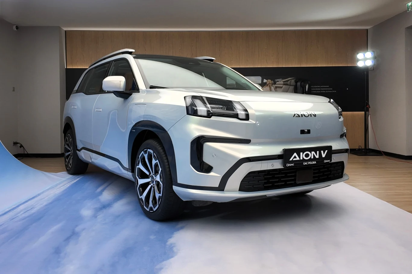 Biały samochód elektryczny Aion V ustawiony w nowoczesnej sali ekspozycyjnej, z drewnianą ścianą w tle i oświetleniem studyjnym, widoczne charakterystyczne reflektory i duże felgi.