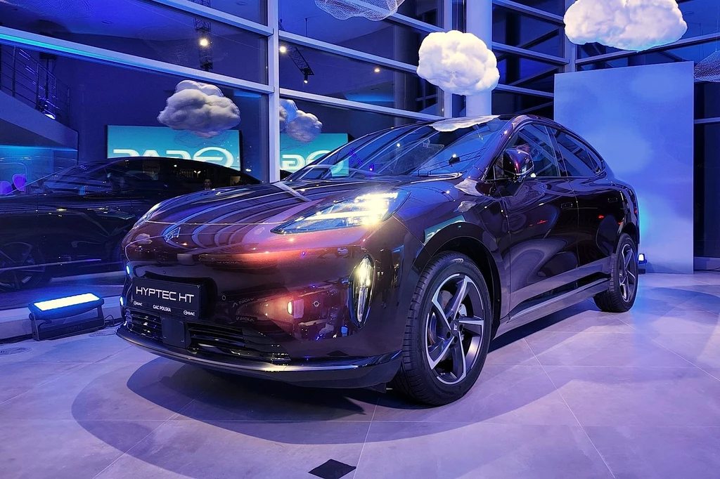 Blisko pięciometrowy Hyptec HT pod kątem rozmiarów plasuje się w grupie takich samochodów jak BMW iX. Czarny samochód elektryczny o nowoczesnym designie prezentowany w oświetlonym wnętrzu z dużymi szybami, w otoczeniu sztucznych chmur, stojący na wystawie motoryzacyjnej.