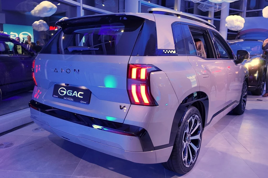 Aion V może przejechać na jednym ładowaniu do 510 km w cyklu mieszanym. Nowoczesny SUV w jasnym kolorze wystawiony w salonie samochodowym z charakterystycznymi światłami tylnymi i dużymi felgami, otoczony nastrojowym oświetleniem wnętrza.