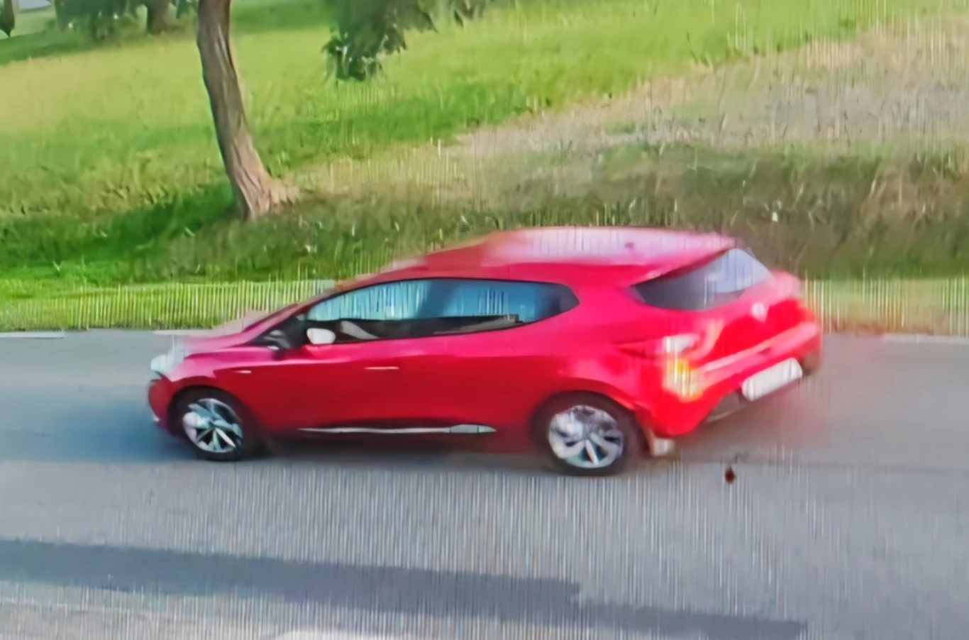 Policja poszukuje czerwonego Renault Clio. Czerwony samochód osobowy poruszający się po drodze asfaltowej, w tle zielona trawa i drzewa.