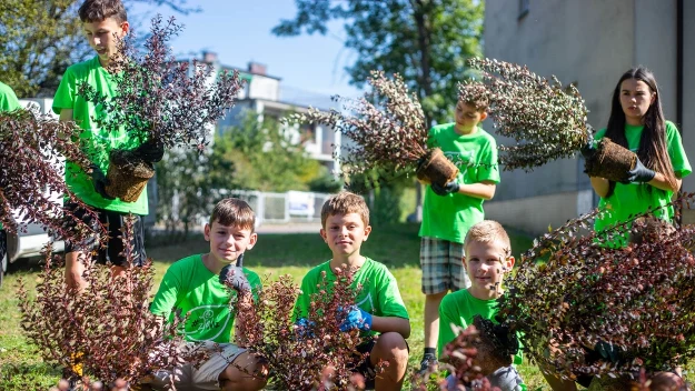 Grupa dzieci w zielonych koszulkach uczestniczy w akcji sadzenia krzewów, każde dziecko trzyma doniczkę z rośliną, a wokół panuje słoneczna, letnia atmosfera. Budynki i drzewa widoczne w tle podkreślają miejskie otoczenie.