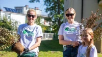 Trzy osoby ubrane w białe koszulki z logo 'Czyste Zielone Miasta', dwie dorosłe kobiety trzymające sadzonki drzew oraz dziewczynka stojąca obok nich na tle zielonego trawnika i miejskich zabudowań.
