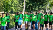 Grupa dzieci i młodzieży w zielonych koszulkach z nadrukiem idzie ścieżką w parku, trzymając śmieci w workach oraz tabliczkę, wokół nich bujna, zielona roślinność.