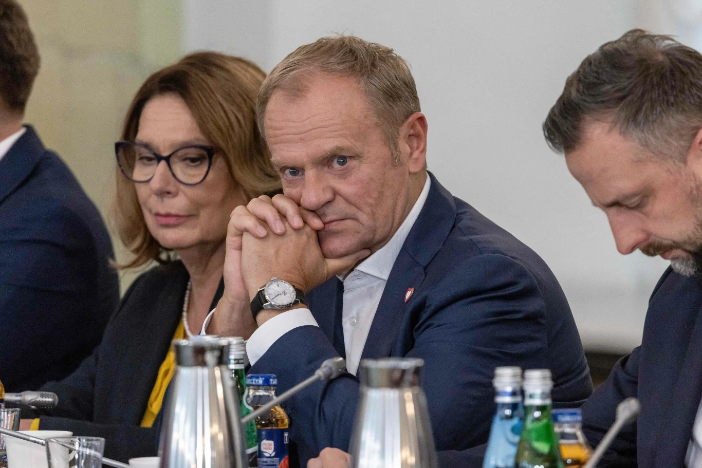 Donald Tusk odpowiedział prezydentowi USA ws. dronów Troje poważnych dorosłych siedzi przy stole konferencyjnym, w otoczeniu butelek wody mineralnej oraz karafek, skupionych na rozmowie lub rozważaniach, w tle neutralne wnętrze sali spotkań.