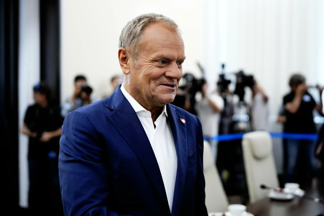 Premier Donald Tusk Mężczyzna w granatowej marynarce i białej koszuli stoi w jasnym pomieszczeniu konferencyjnym, w tle grupa fotoreporterów z aparatami, widoczne krzesła i filiżanki na stole.