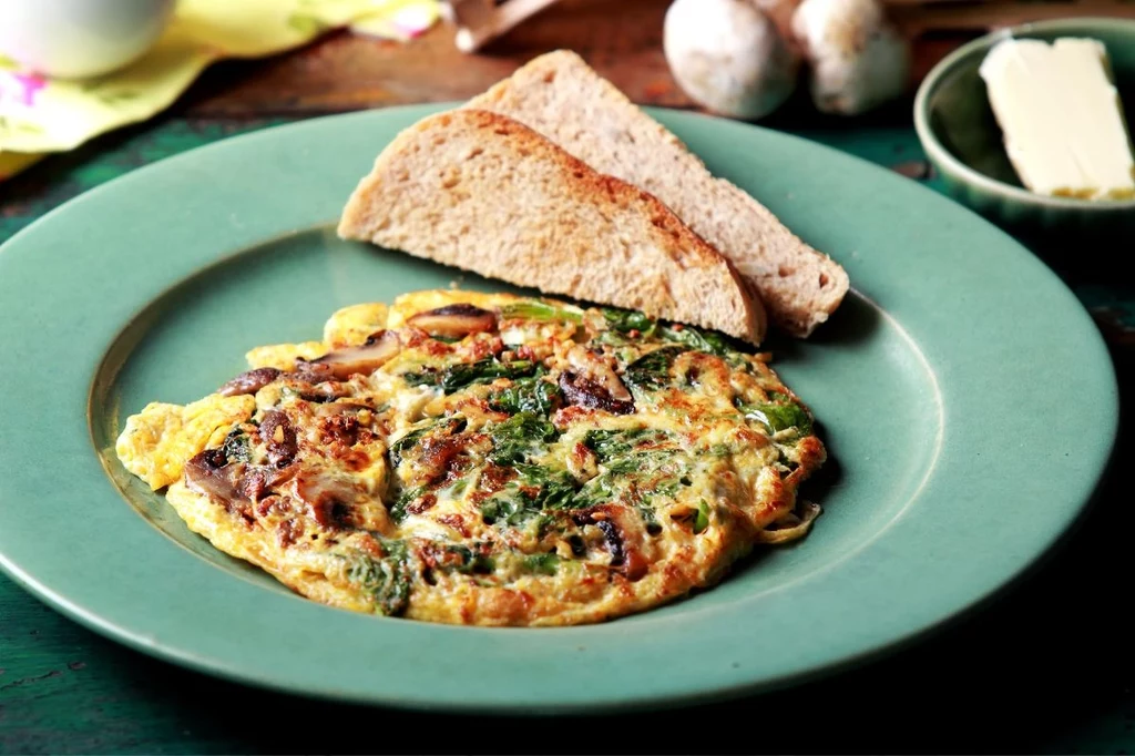 Omlet z warzywami i ziołami podany na zielonym talerzu, obok dwie kromki chleba, w tle masło na małym talerzyku oraz kilka grzybów.
