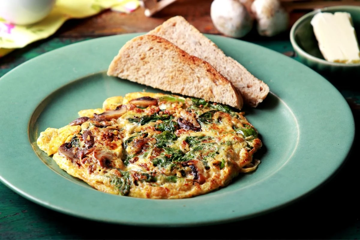 Omlet z warzywami i ziołami podany na zielonym talerzu, obok dwie kromki chleba, w tle masło na małym talerzyku oraz kilka grzybów.