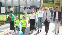 Grupa dzieci w kamizelkach odblaskowych i dorośli idą chodnikiem przed budynkiem szkoły, dzieci trzymają worki na śmieci, sugerując udział w akcji sprzątania.