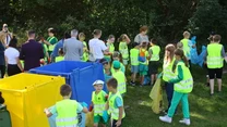 Dzieci w odblaskowych kamizelkach segregują śmieci podczas zajęć terenowych, wokół nich ustawione są kolorowe pojemniki na odpady, a w tle widoczni są opiekunowie.