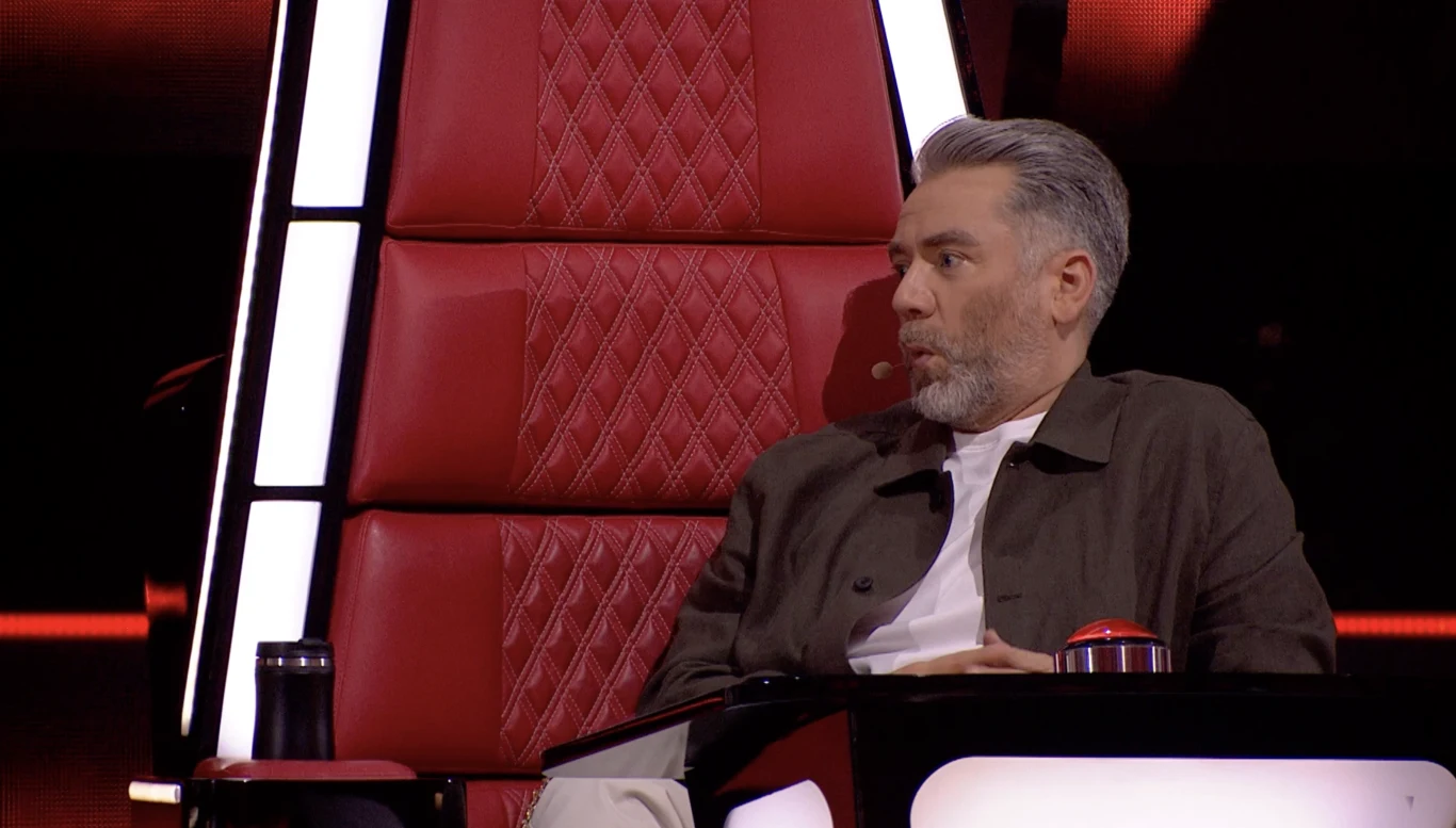 "The Voice of Poland": Kuba Badach nie mógł wyjść z podziwu po występie 27-latka Sędzia programu telewizyjnego siedzący w charakterystycznym czerwonym fotelu z mikrofonem przy twarzy, obok przycisk i kubek, w tle ciemne oświetlenie sceniczne.