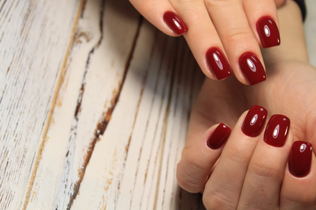 Dłonie z eleganckim manicure, paznokcie pomalowane lakierem w ciemnoczerwonym kolorze na tle bielonego, drewnianego stołu.