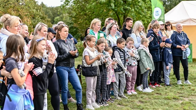 Grupa ludzi, w tym dzieci i dorośli, stoi na trawie w parku, trzymając w rękach drobne przedmioty i ubrana w codzienne, wygodne ubrania, niektórzy klaszczą, wszyscy zwróceni są w jednym kierunku jakby obserwowali występ lub wydarzenie, w tle drzewa i n...