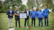 Grupa sześciu mężczyzn stoi na trawniku wokół świeżo posadzonego drzewa, trzymają tablicę z napisem promującym inicjatywę ekologiczną. W tle widoczne drzewa oraz fragment białego namiotu, dający wrażenie wydarzenia plenerowego.