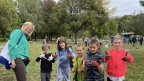 Grupa uśmiechniętych dzieci w rękawiczkach i kobieta stoją na trawniku w parku, dzieci pokazują brudne dłonie po pracy w ziemi, w tle widać innych uczestników i drzewa.