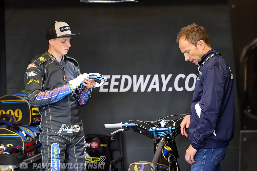 Młody żużlowiec w kombinezonie sportowym przygotowuje rękawice przed motocyklem żużlowym, obok stoi dorosły mężczyzna w sportowej kurtce, obaj skupieni na przygotowaniu sprzętu do wyścigu.