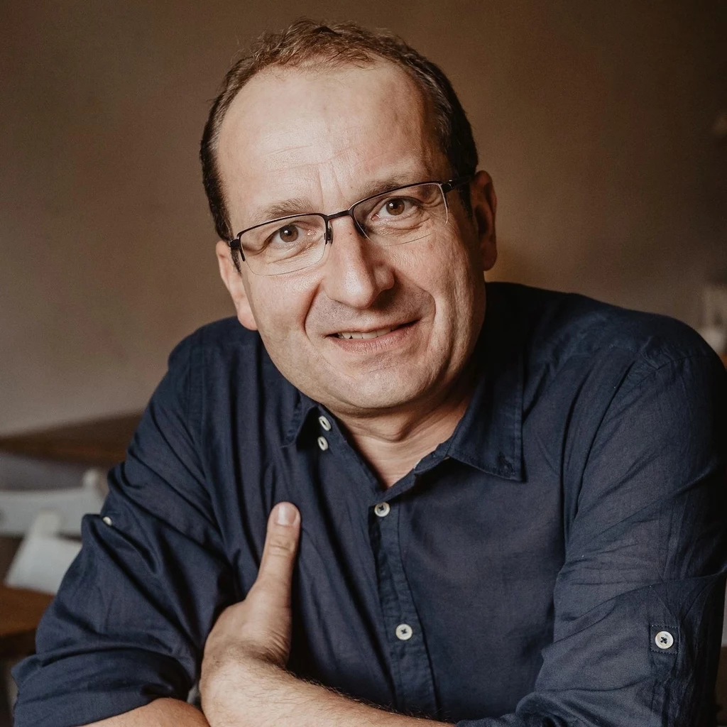 Robert Górski Mężczyzna w średnim wieku w okularach siedzi przy stole z lekko uśmiechniętą twarzą, ubrany w granatową koszulę, w tle rozmyte wnętrze pomieszczenia.