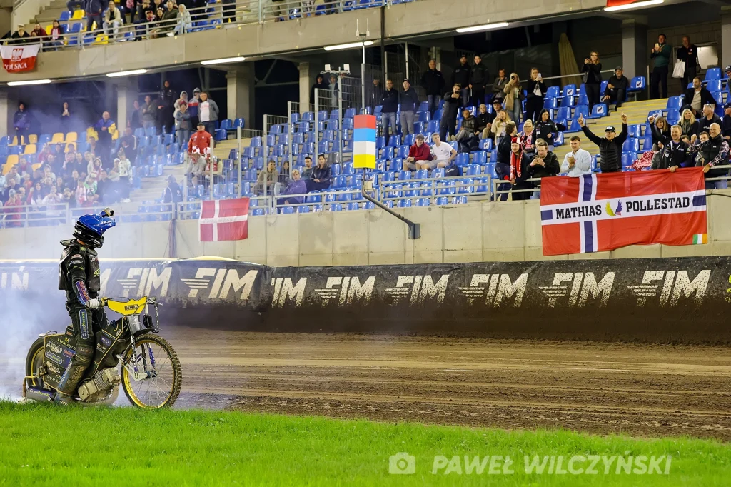 Żużlowiec w pełnym stroju ochronnym i kasku stoi na torze pośród unoszącego się dymu, na trybunach widać kibiców, flagi narodowe oraz transparent z nazwiskiem zawodnika.