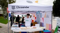 Stoisko promocyjne pod białym namiotem z napisem 'Chrzanów', na stole leżą materiały informacyjne, torby i inne przedmioty promocyjne, za stołem stoi młoda osoba w białym fartuchu, wokół widoczne są inne stoiska oraz odwiedzający.