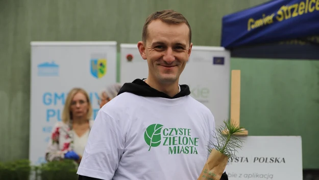 Uśmiechnięty mężczyzna ubrany w biały t-shirt z napisem 'Czyste Zielone Miasta' trzyma w rękach małą sadzonkę drzewka. W tle widoczny jest namiot oraz osoby uczestniczące w wydarzeniu.