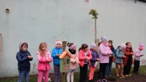 Grupa dzieci w kolorowych kurtkach stoi w rzędzie przy ścianie budynku na zewnątrz, obok nowo posadzonego drzewa i domku dla owadów.