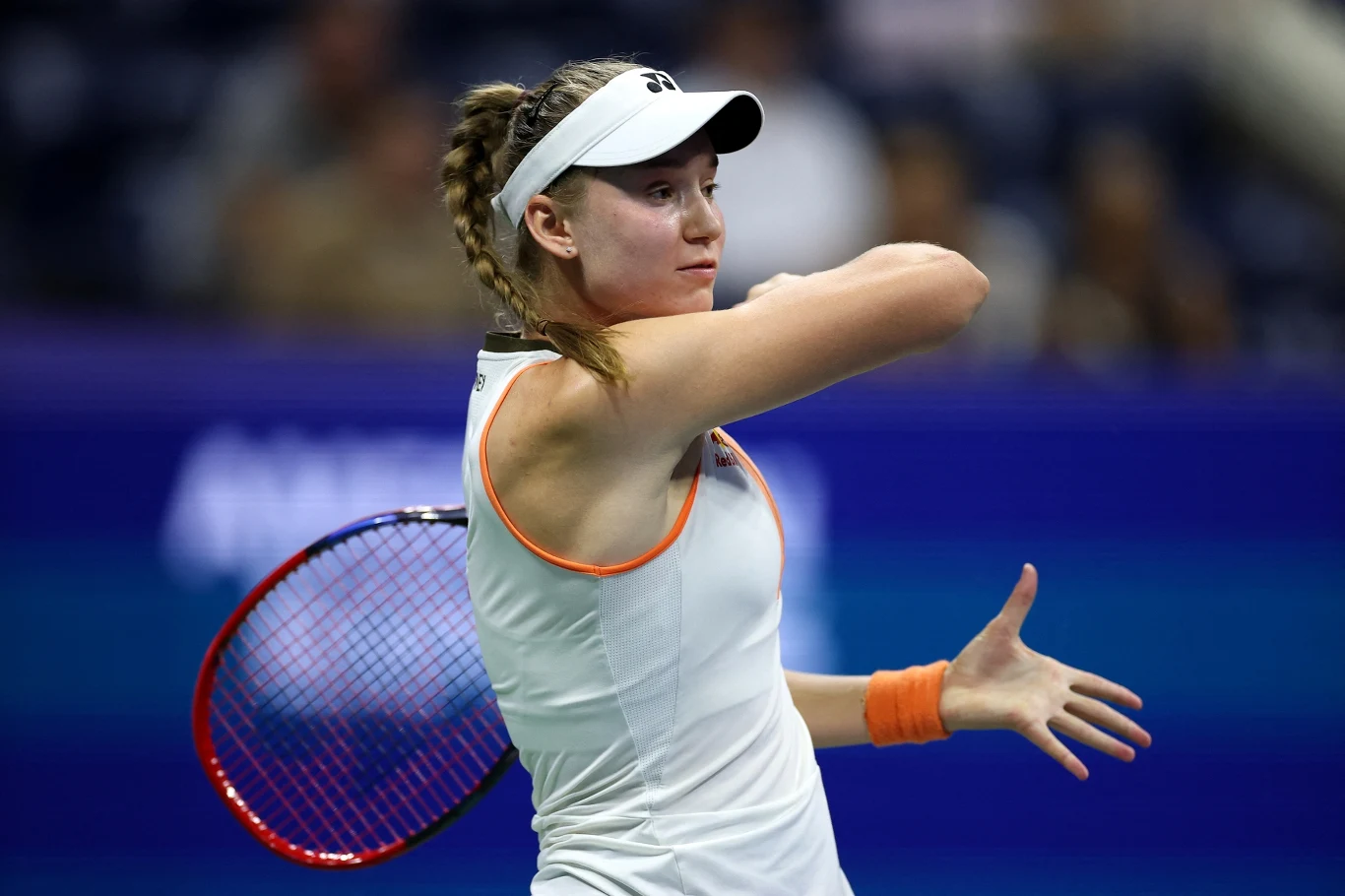 Jelena Rybakina Tenisistka w białym stroju i białej czapce wykonuje zamach rakietą podczas meczu, w tle rozmazane trybuny i niebieski kort.