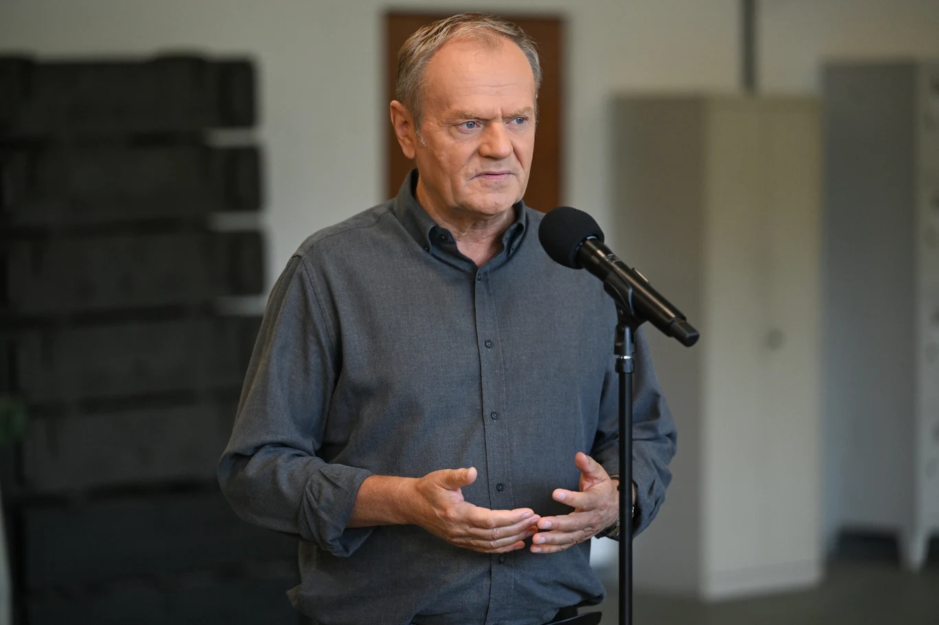 Tusk z wizytą w zakładach produkcji amunicji. Tyle pocisków powstanie w rok Mężczyzna w średnim wieku w szarej koszuli mówi do mikrofonu ustawionego na stojaku, stojąc w jasnym, nowoczesnym wnętrzu