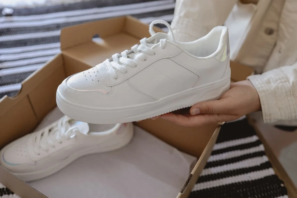 Nienoszone, nowe buty symbolizują zatrzymanie się w drodze. Lepiej oddać je lub odsprzedać Biała, skórzana tenisówka trzymana w ręce nad otwartym pudełkiem, drugi but spoczywa w środku pudełka na papierze.