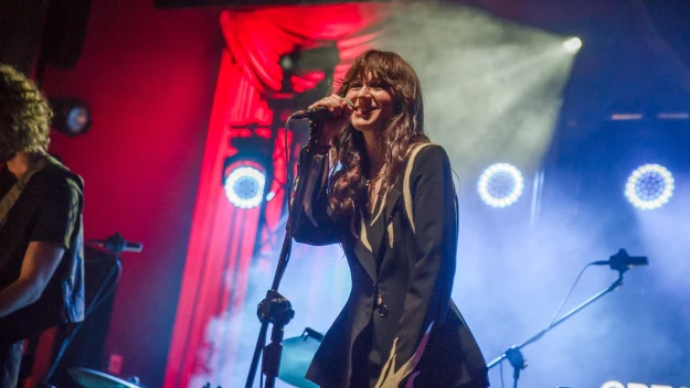 Kobieta śpiewa do mikrofonu na scenie koncertowej, za nią widoczne są kolorowe światła i elementy perkusji, panuje atmosfera energetycznego występu muzycznego.