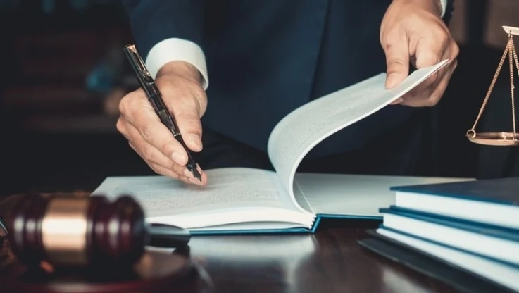 Mężczyzna w garniturze podpisuje dokumenty na biurku, obok leży drewniany młotek sędziowski oraz waga symbolizująca sprawiedliwość, stos papierów stanowi tło.