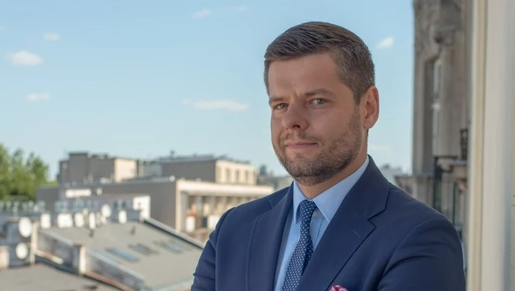 Mężczyzna w garniturze i krawacie stoi na tle miejskiej panoramy, z lekko poważnym wyrazem twarzy, przy jasnym dziennym świetle, w tle widoczne są nowoczesne budynki i fragment fasady kamienicy.
