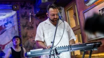 Mężczyzna w białej koszuli grający na klawiaturze elektronicznej na scenie, za nim perkusista w ciemnym podkoszulku wykonuje swoją partię, całość oświetlona kolorowymi światłami w klubowym wnętrzu.