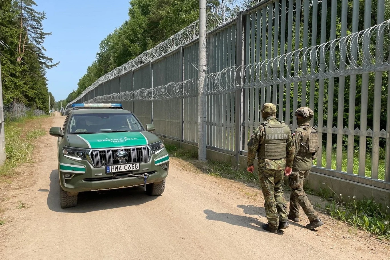 Zatrzymano Afgańczyka, który miał zaatkować polskiego żołnierza i przemycać migrantów dwóch funkcjonariuszy ubranych w mundury patroluje teren wzdłuż wysokiego ogrodzenia z drutem kolczastym, obok zaparkowany samochód służbowy oznakowany symbolem straży granicznej, po obu stronach droga żwirowa, w tle las