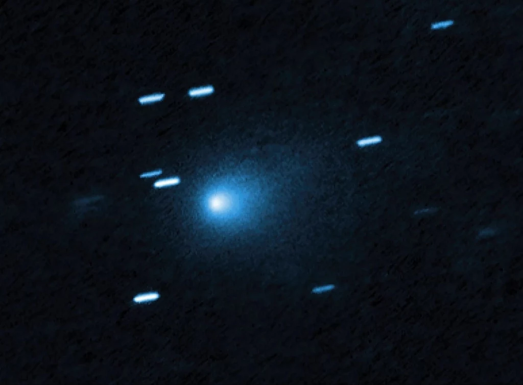 Jasny obiekt w centrum otoczony rozmytą poświatą, widoczny na tle ciemnego nieba z kilkoma rozmazanymi śladami światła wskazującymi na ruch innych obiektów astronomicznych.