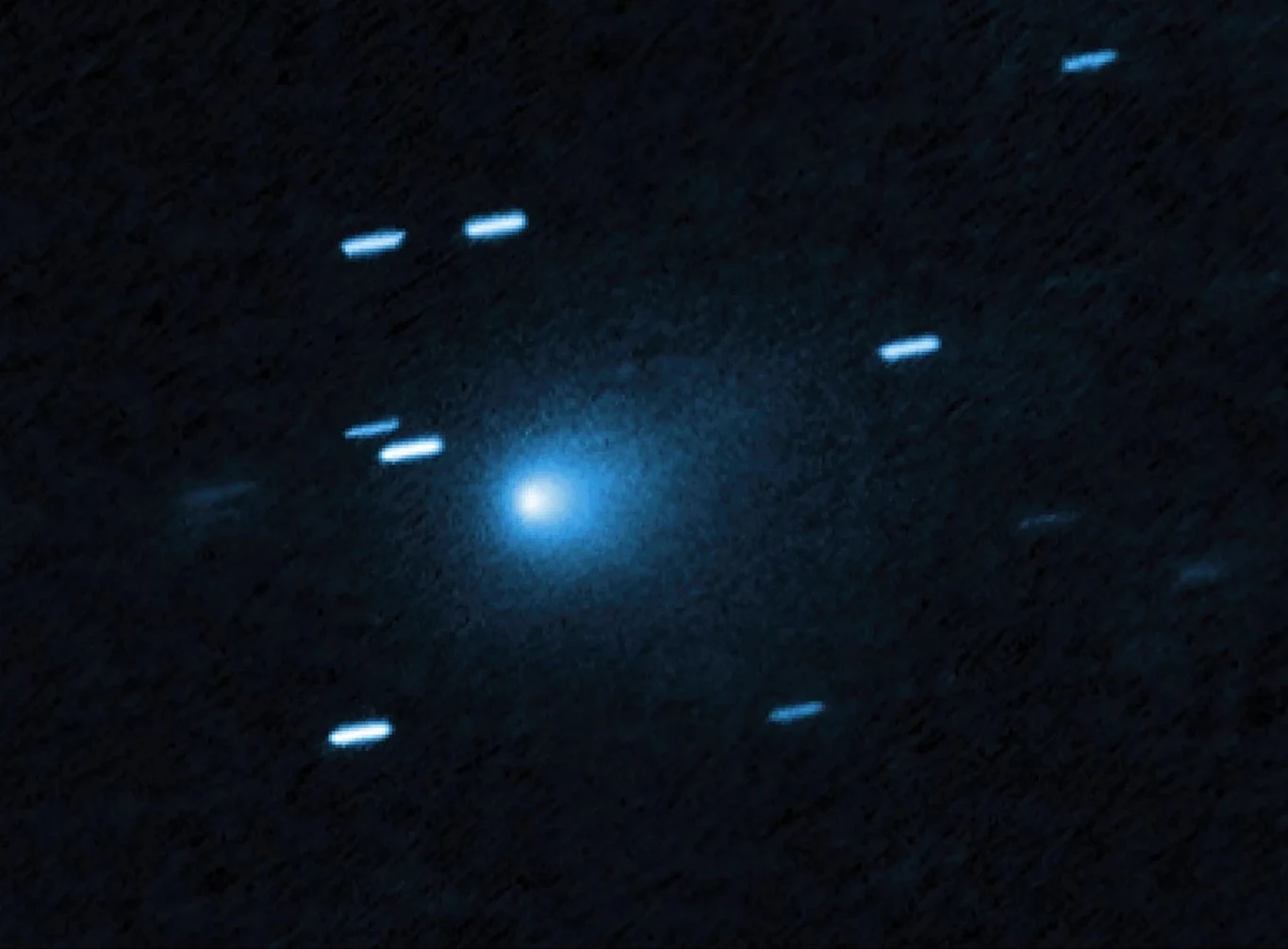 Kometa 3I/ATLAS uchwycona przez Kosmiczny Teleskop Hubble'a. Jasny obiekt w centrum otoczony rozmytą poświatą, widoczny na tle ciemnego nieba z kilkoma rozmazanymi śladami światła wskazującymi na ruch innych obiektów astronomicznych.