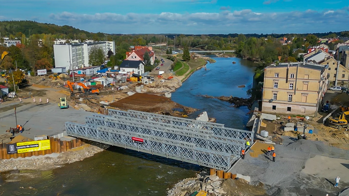 Rok temu południowo-zachodnią Polskę nawiedziła powódź. Na odbudowę zniszczeń w obszarze infrastruktury zmobilizowano już ponad 1 mld zł Metalowy most tymczasowy rozciąga się nad rzeką w centrum miasta, wokół widoczne są maszyny budowlane, robotnicy w pomarańczowych ubraniach oraz zabudowania miejskie, w tle rozpościera się zielony krajobraz