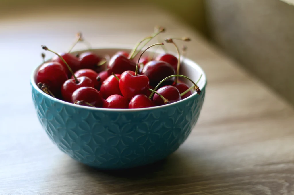 Odmiana szarlotki wykorzystująca regionalne grójeckie jabłka oraz wiśnie Miseczka z jasnoniebieskiej ceramiki wypełniona świeżymi, czerwonymi czereśniami ze szypułkami stoi na drewnianym blacie.