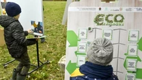 Dwoje dzieci bawi się na świeżym powietrzu przy dużej planszy z ekologicznymi zagadkami, ubrani są w jesienne ubrania, wokół trawa i liście.