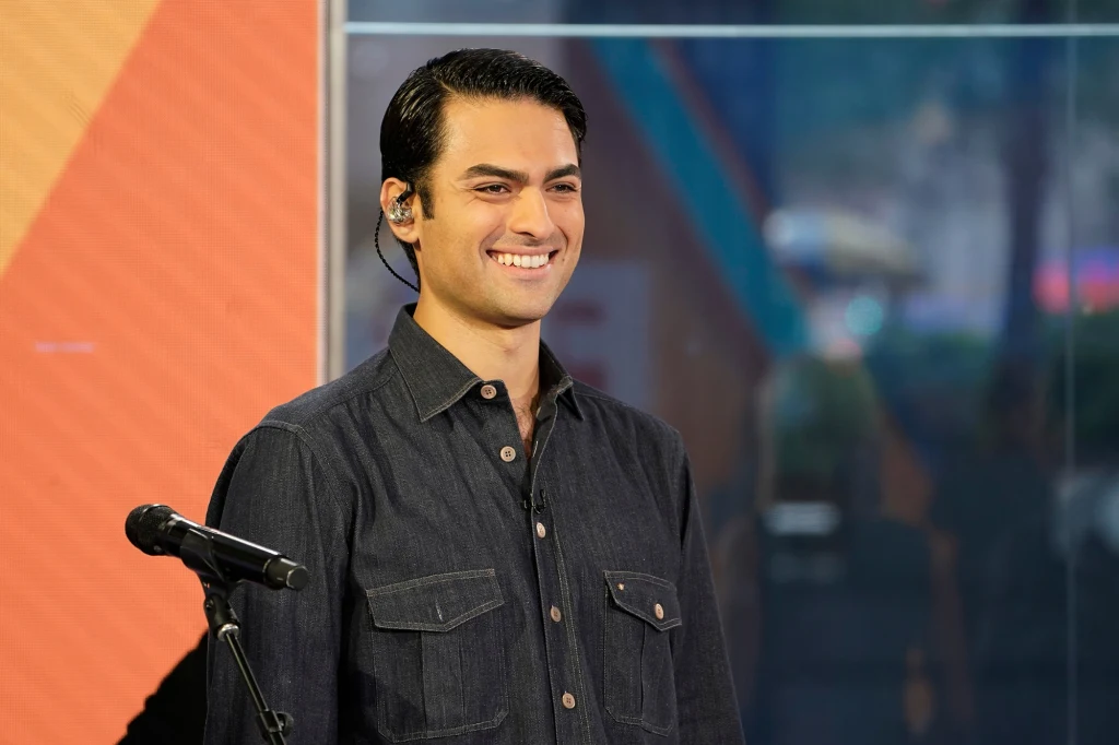 Uśmiechnięty młody mężczyzna w ciemnej koszuli stoi przed mikrofonem na tle kolorowej ściany i przeszklonego panelu.