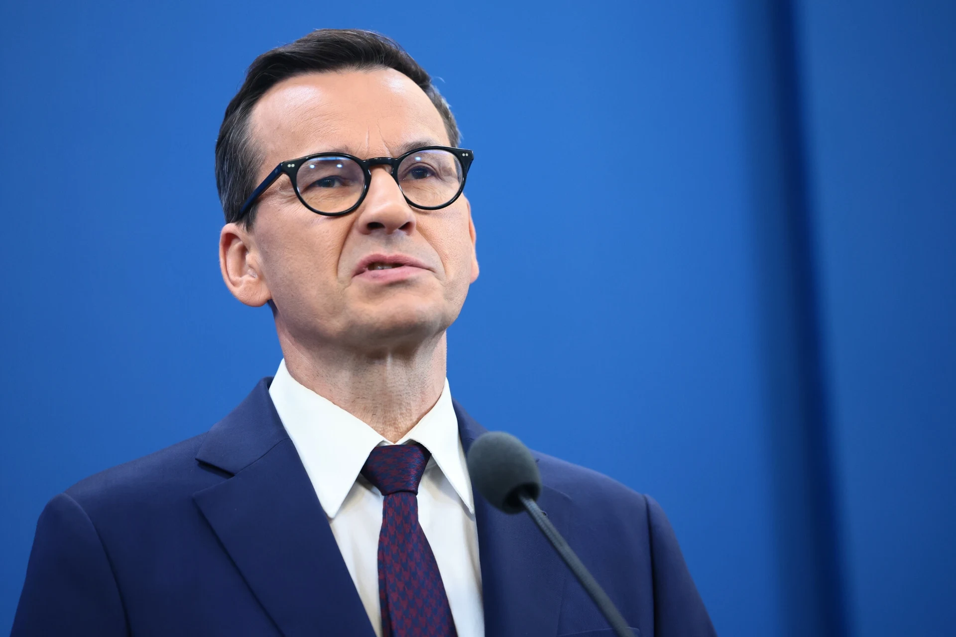 Mężczyzna w garniturze i okularach przemawia do mikrofonu na tle niebieskiego ekranu.