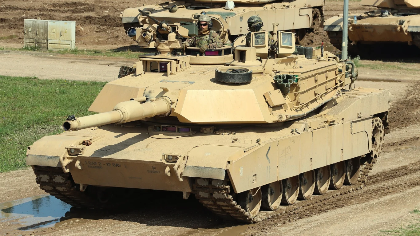 Czołg M1A2 SEPv3 Abrams Amerykański czołg Abrams jadący po polu, prowadzony przez kilku żołnierzy w mundurach, z otwartymi włazami i widocznym uzbrojeniem.