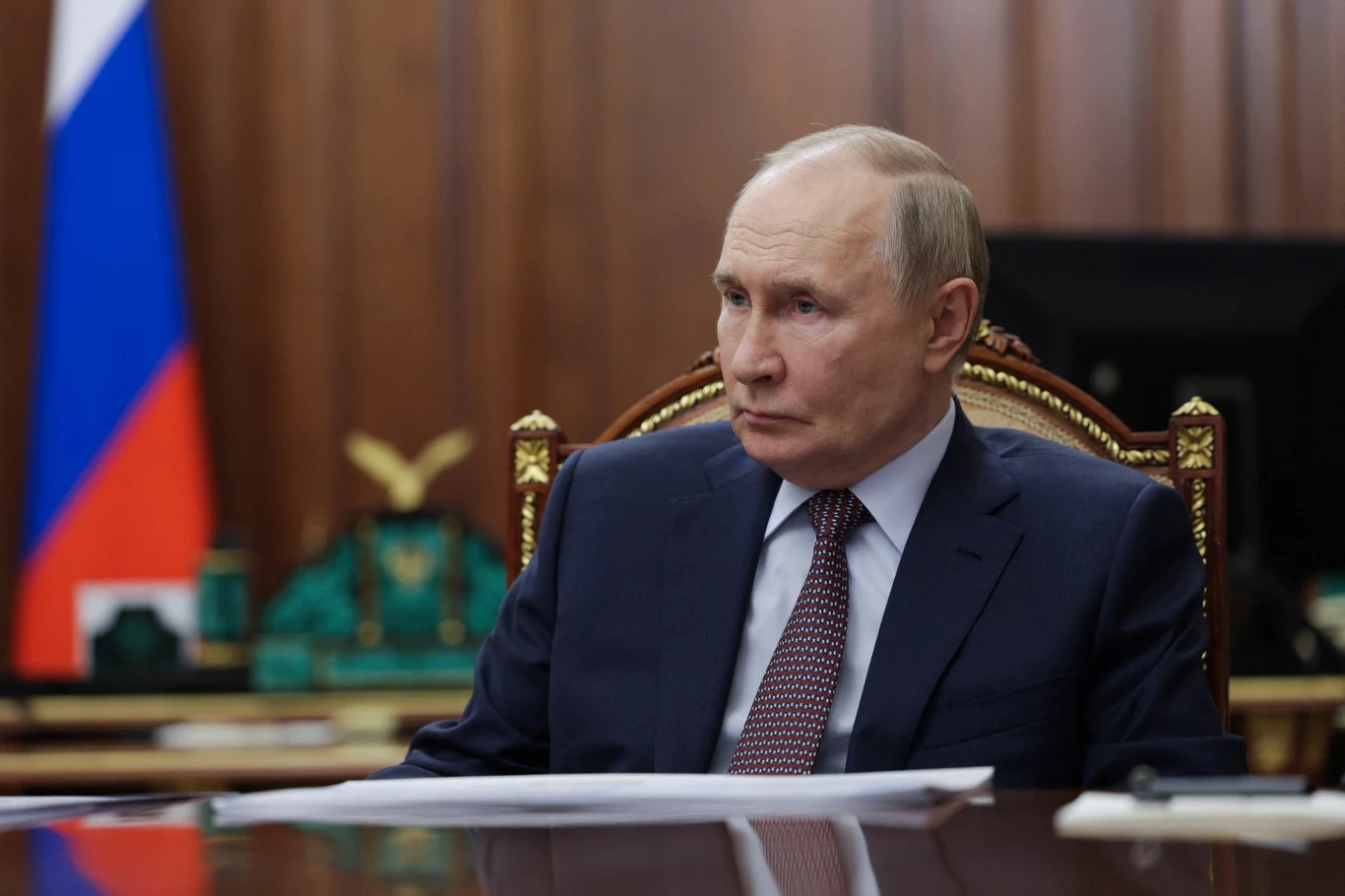 KE: Rosja musi zapłacić za swoje zniszczenia, Moskwa zapowiada konkretne kroki (zdj. ilustracyjne) Mężczyzna w garniturze siedzący przy dużym, ozdobnym biurku na tle rosyjskiej flagi i ciemnego, drewnianego wnętrza.