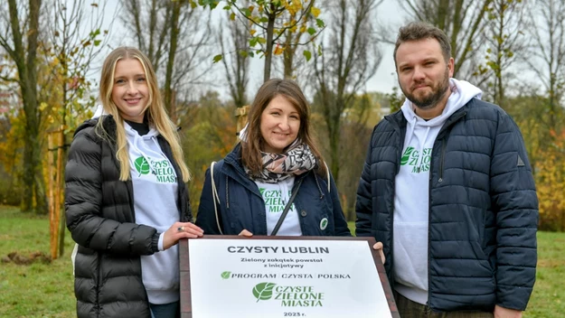 Troje uśmiechniętych dorosłych stoi na zewnątrz na tle jesiennych drzew, trzymając tablicę informacyjną dotyczącą programu ekologicznego „Czyste Zielone Miasta” w Lublinie.