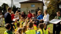 Grupa dzieci w odblaskowych kamizelkach zgromadzona na świeżym powietrzu przed budynkiem, otoczona przez dorosłych wręczających im przedmioty, panuje radosna atmosfera podczas działania o charakterze edukacyjnym.