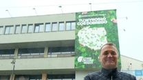 Mężczyzna w ciemnej kurtce stoi na tle budynku, na którym widnieje duży baner z zielonym motywem oraz mapą Polski i napisem 'Tu wyrosło coś dobrego'.
