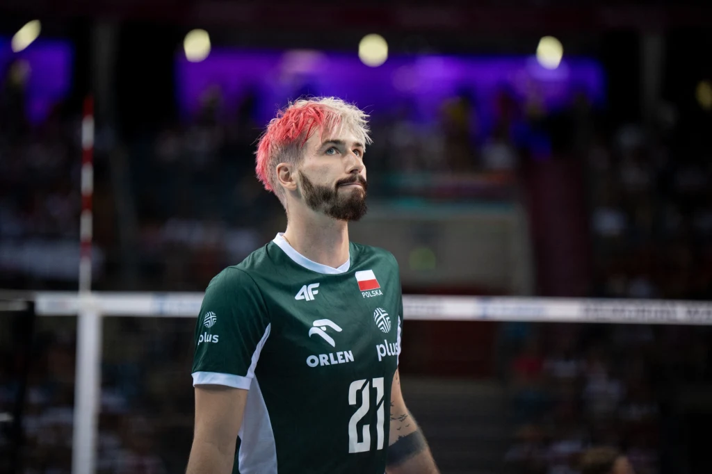 Siatkarz ubrany w zielony strój sportowy z numerem 21 oraz emblematami sponsorów i flagą Polski, wyróżniający się charakterystycznie zafarbowanymi włosami, znajduje się na hali sportowej przy siatce, w tle rozmyte trybuny z widownią.