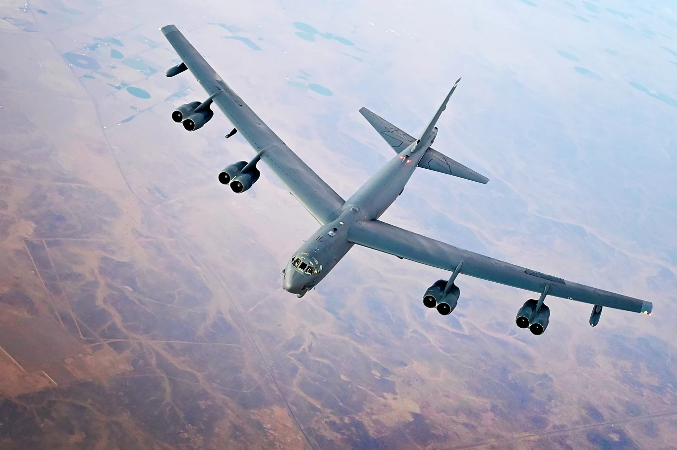 Bombowiec strategiczny B-52H z 5. Skrzydła Bombowego Duży bombowiec strategiczny z ośmioma silnikami, lecący nad suchym, rozległym terenem, widok z powietrza z wyraźnie widoczną sylwetką samolotu i krajobrazem poniżej.